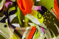Close-up image of 100% silk square scarf featuring a collage of fruits, vegetables, and flowers in luscious shades of green and yellow, with pops of orange and pink throughout, illustrating the lightly ridged texture of the silk twill along with the rich color tones and luminous nature of the silk scarf.
