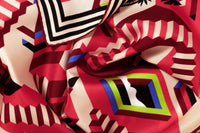 Close-up image of 100% silk square scarf featuring a motif of interlocking staircases and platforms in various shades of red accented with pops of blue and green. A neutral stripe border around the scarves' edges. Illustrates the lightly ridged texture of the silk twill along with the rich color tones and luminous nature of the silk scarf.