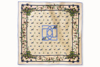 Flatlay image of 100% silk square scarf featuring an English garden-inspired floral illustration motif with a periwinkle blue version of the brand logo dominantly featured in the middle of the design against a warm neutral background and a periwinkle blue edge.