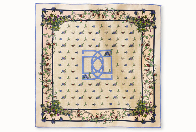Flatlay image of 100% silk square scarf featuring an English garden-inspired floral illustration motif with a periwinkle blue version of the brand logo dominantly featured in the middle of the design against a warm neutral background and a periwinkle blue edge.