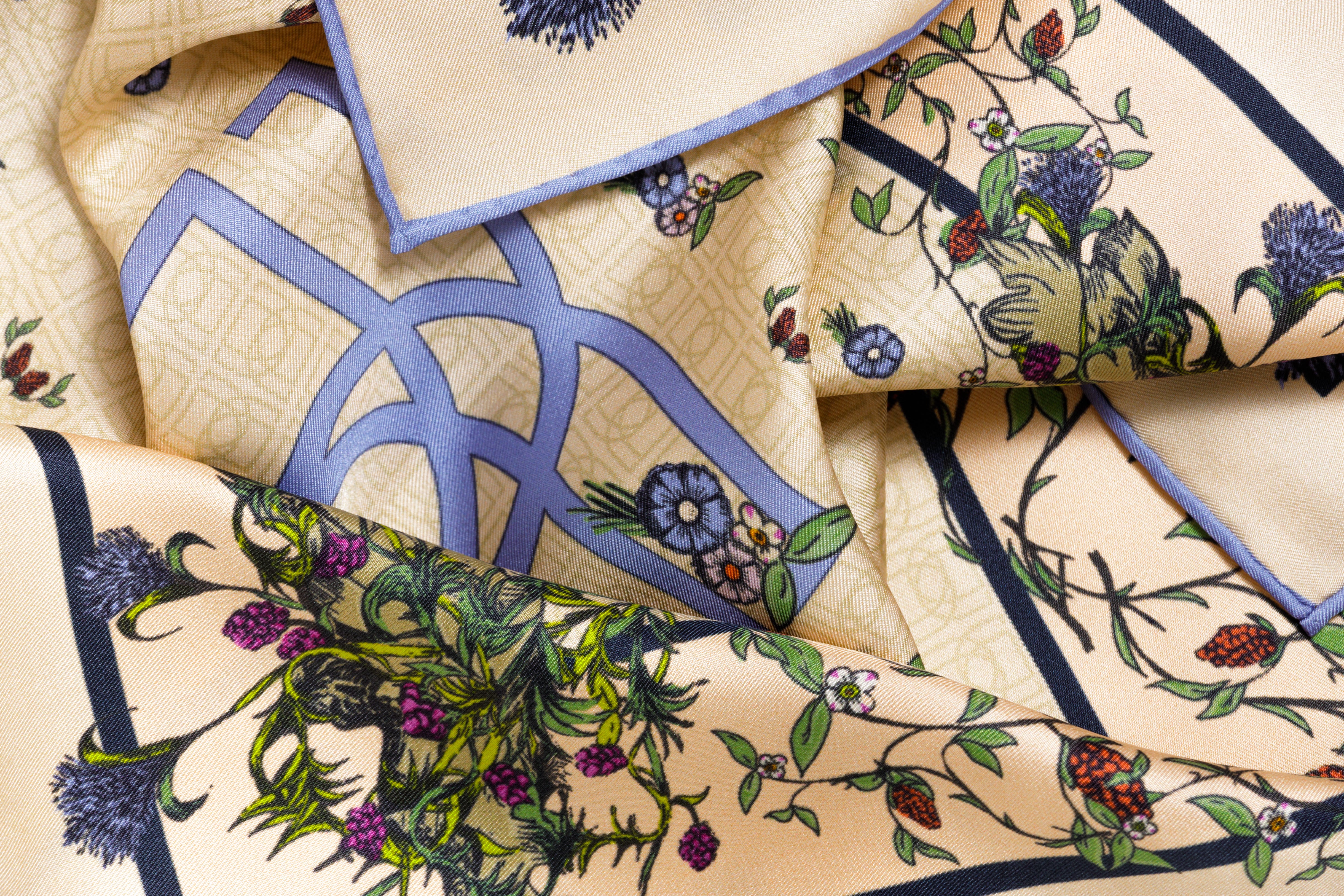 Close-up image of 100% silk square scarf featuring an English garden-inspired floral illustration motif with a periwinkle blue version of the brand logo dominantly featured. Illustrates the lightly ridged texture of the silk twill along with the rich color tones and luminous nature of the silk scarf.