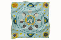 Flatlay image of 100% silk square scarf featuring a motif of stars, the sun and the moon phases positioned around planet Earth. Details in shades of rich turquoise, marine blue, white, chartreuse and maize on a light turquoise background.