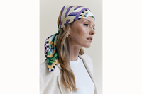 Image of a female model wearing a scarf wrapped around her head, tied in the back, styled as a headscarf. Model is wearing a white tank top and a cream blazer.