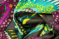 Close-up image of 100% silk square scarf featuring a motif of two large-scale cranes with brightly colored wings in shades of blue and green with geometric designs. The scarf’s center background is a tonal army green and the border features irregular grey polka dots on a magenta background. Illustrates the lightly ridged texture of the silk twill along with the rich color tones and luminous nature of the silk scarf.