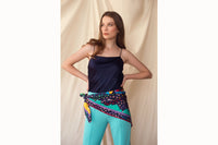 Image of female model standing with hands on hips while wearing the scarf tied around her waist. Model is also wearing a navy tank top and turquoise trousers.