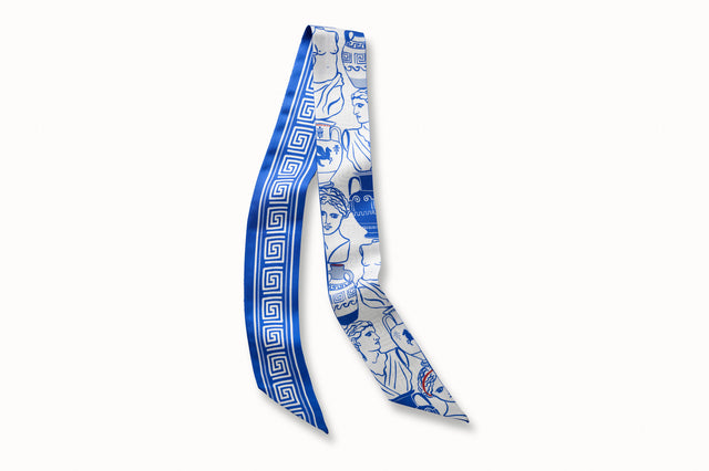 Flatlay image of 100% silk ribbon style scarf, 2” wide by 32” long. Featuring a illustrations of Greek goddess busts and urns in a bright Aegean blue on a white background. The opposite side of the scarf features a white Greek key design motif on a bright blue background.