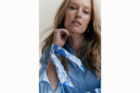 Image of a female model wearing the scarf wrapped around her neck with tail ends of the scarf draping onto her arm. Model is wearing a light blue button down and her hair is brushed to the right side of her body.