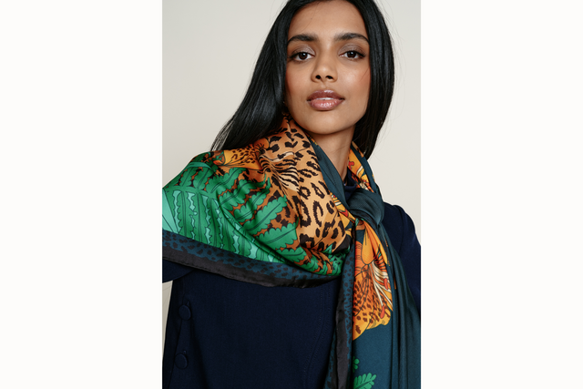 Image of a female model wearing the scarf in a loose knot tied in the front. Model wears a navy blue top and looks directly into the camera