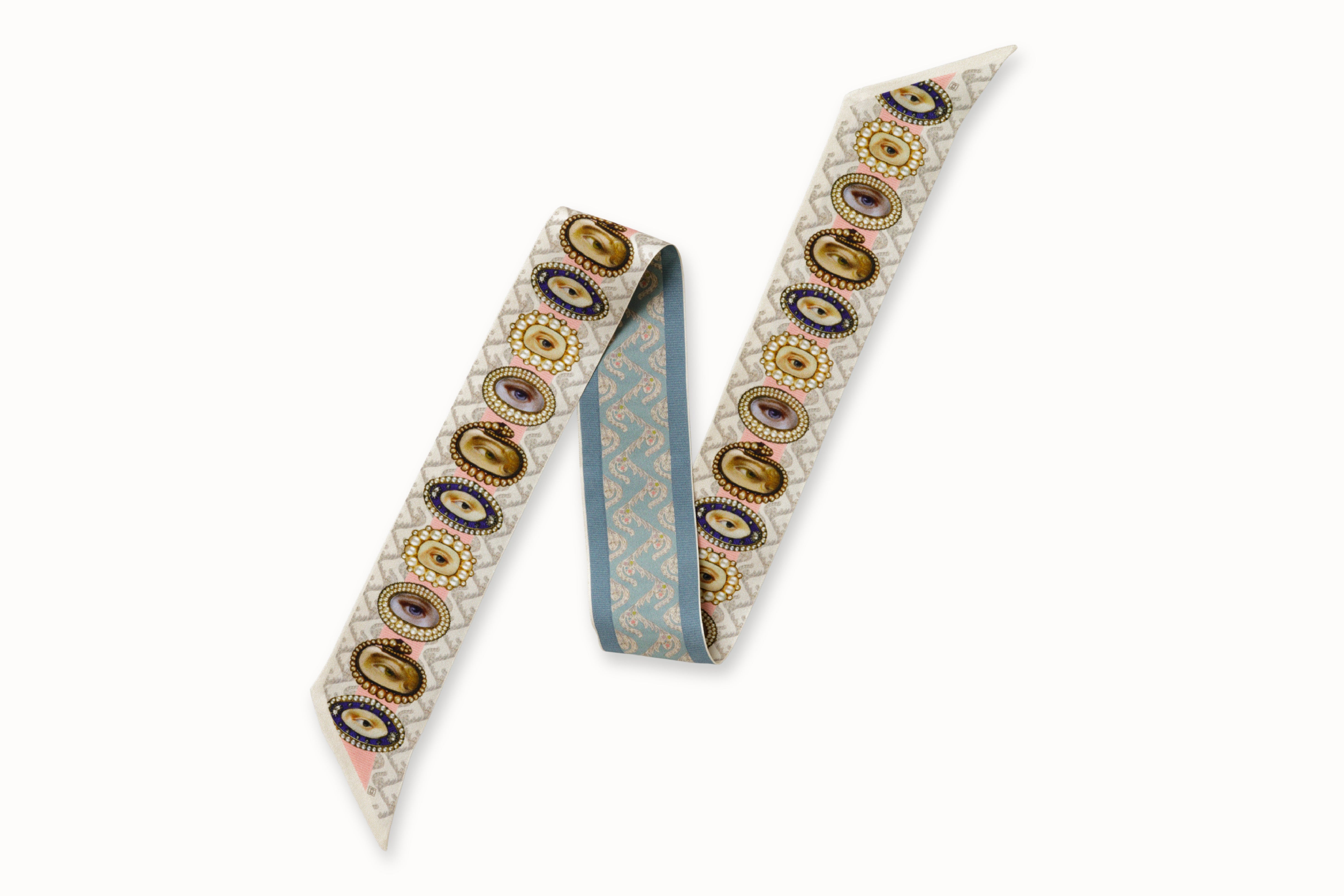 Flatlay image of 100% silk ribbon style scarf, 2” wide by 32” long, folded in a zig-zag shape. Design features a series of hand-painted lover's eye jewels on one side and hand drawn wallpaper design with a floral motif in a soft blue tone on the other side.