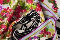 Close-up image of 100% silk square scarf featuring a motif of three black female stick figures dressed for a night out and standing in front of the hotel entrance surrounded by urns and floral arches. The elements are in various shades of pink, green, soft red and baby blue splashed across a white background. Illustrates the lightly ridged texture of the silk twill along with the rich color tones and luminous nature of the silk scarf.