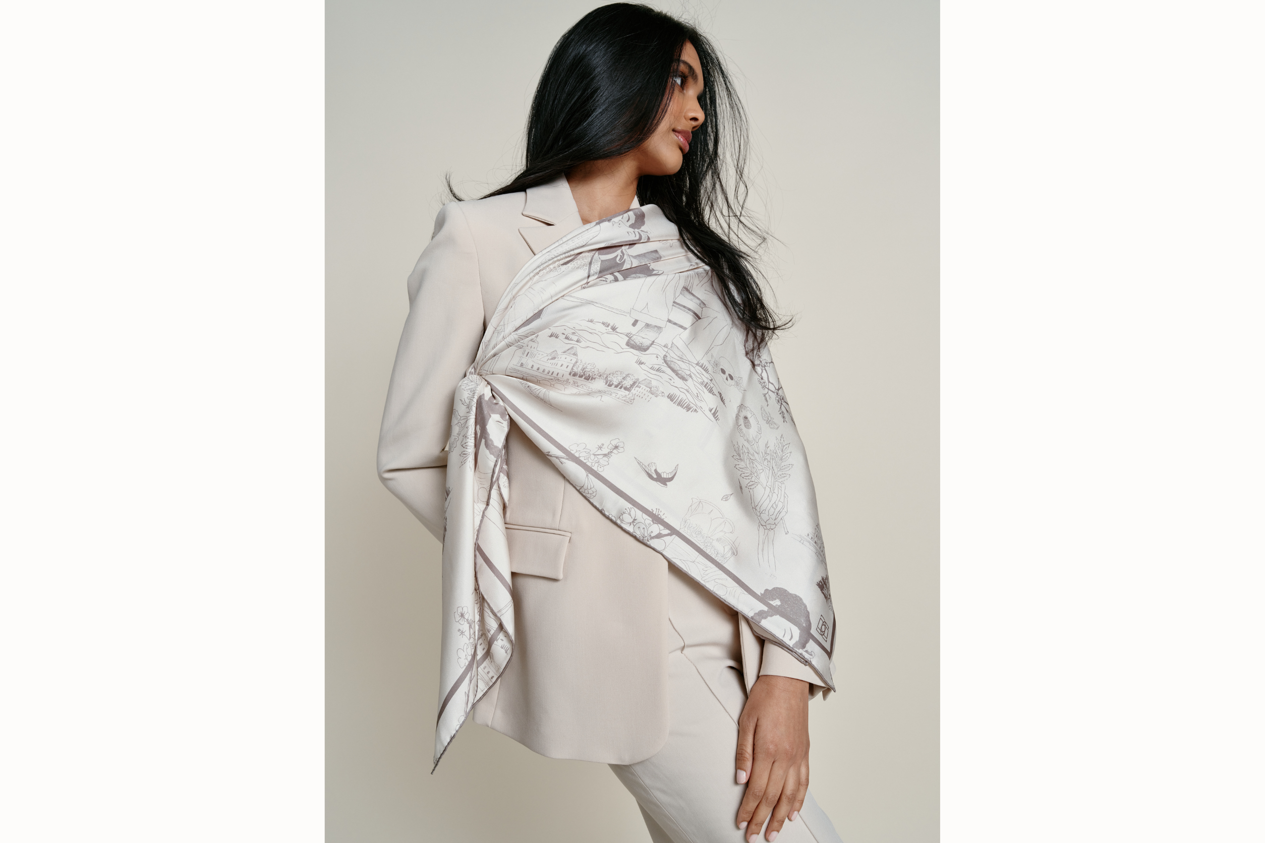 Image of a female model wearing the scarf sashed at the shoulder and tied over a neutral 2-piece blazer set near the waist of the opposite end. Model holds one hand to her slightly popped hip as she looks away from the camera.