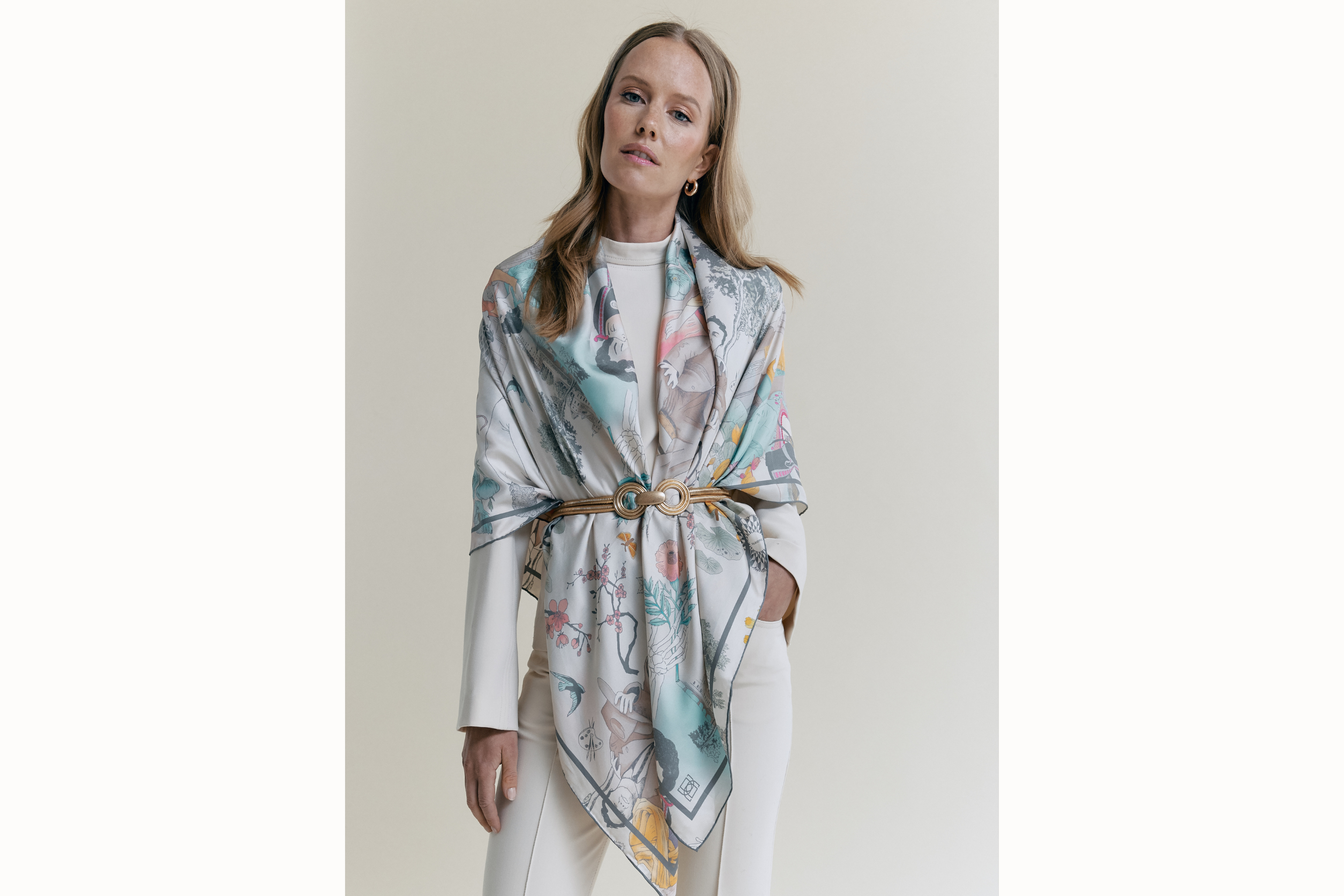 Image of a female model wearing the watercolor version of the scarf over the shoulders and a gold belt secures the scarf at the waist. Model holds her left hand in the pocket of her white pants.