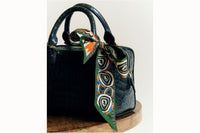 Scarf tied as a bow on the handle of a green small purse which rests on a wooden stool.
