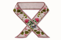 Flatlay image of 100% silk ribbon style scarf, 2” wide by 32” long folded into . Featuring delicate peony flowers + blue butterfly illustrations coupled with a simple soft pink reverse.