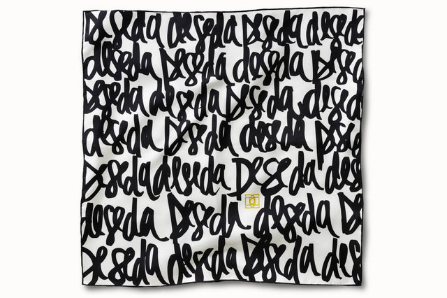 Flatlay image of 100% silk square scarf featuring DESEDA written in black script on a repeat throughout the scarf on a white background. Features one small citron-colored DESEDA logo placed off-center in the design. Thin black border around the scarf’s edges.