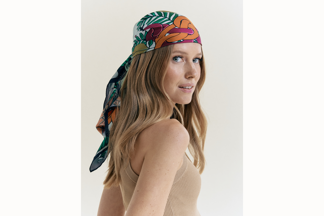 Female model wearing scarf styled as a headscarf and tied at the back of her head. She wears a neutral colored tank and looks over her shoulder into the camera. .