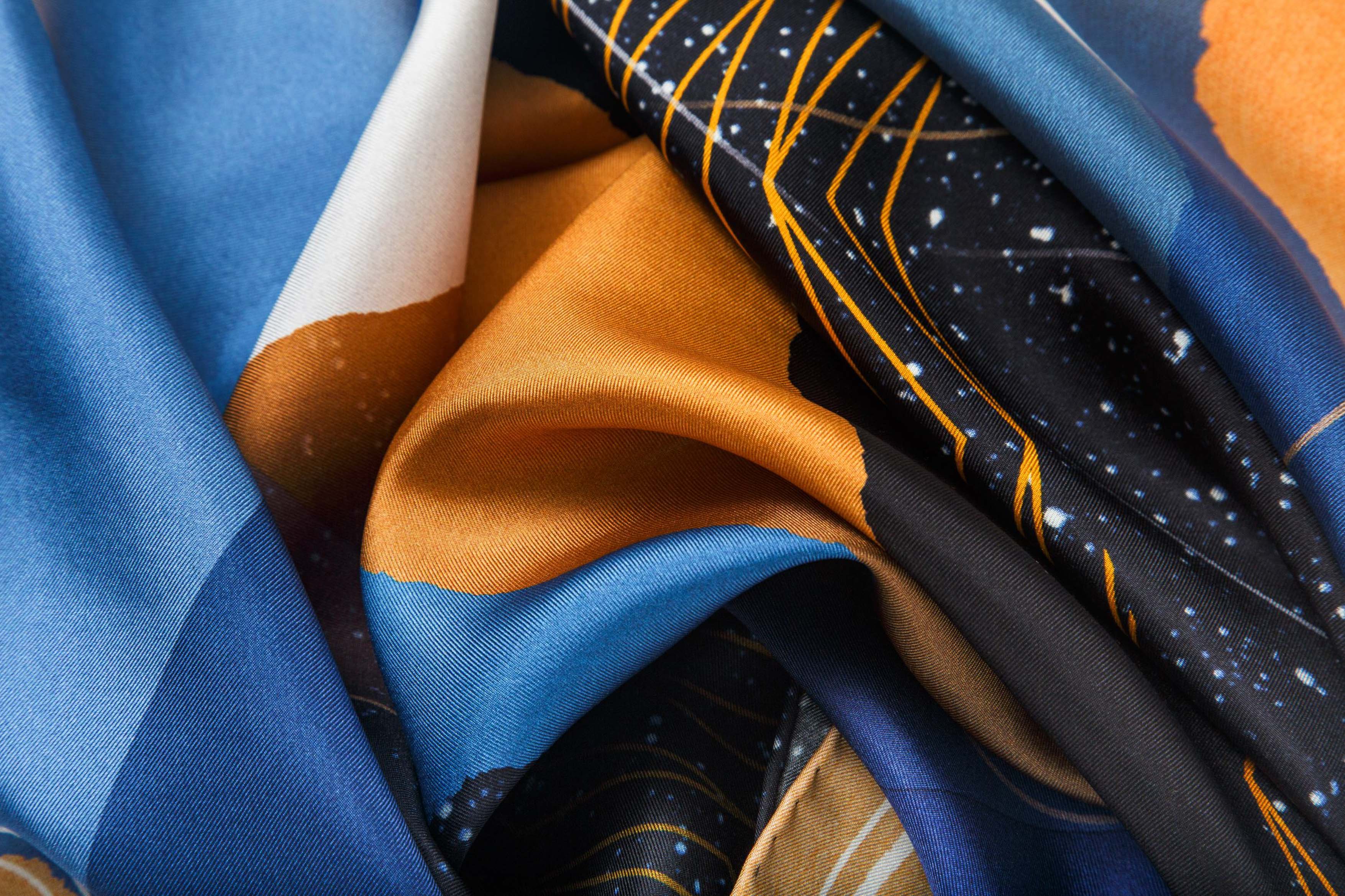Close-up image of 100% silk square scarf featuring a cosmic collage motif, including a large planet in the center surrounded by the moon phases and stars in outer space. Colors used include shades of warm oranges and blues as well as white and black details. Illustrates the lightly ridged texture of the silk twill along with the rich color tones and luminous nature of the silk scarf.
