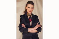Image of female model wearing the scarf tied around the base of her neck and secured with a simple knot. The tail ends of the scarf drape down her chest. Model is also wearing a black blazer.