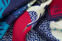Close-up image of 100% silk square scarf featuring an illustration of large crashing waves against a blue and white patterned background with small lighthouse and two cranes flying in front of the moon. Colors include various shades of blue, from deep navy to royal to French blue, as well as warm red and cream. Illustrates the lightly ridged texture of the silk twill along with the rich color tones and luminous nature of the silk scarf.