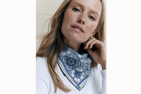Image of the female model wearing a white long-sleeve shirt and the Azulejos scarf tied behind her neck in a bandana style. The model places her hand against her cheek and looks at the camera.