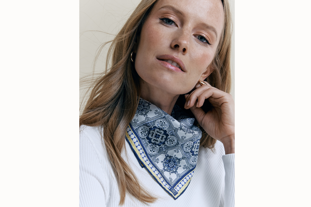 Image of the female model wearing a white long-sleeve shirt and the Azulejos scarf tied behind her neck in a bandana style. The model places her hand against her cheek and looks at the camera.
