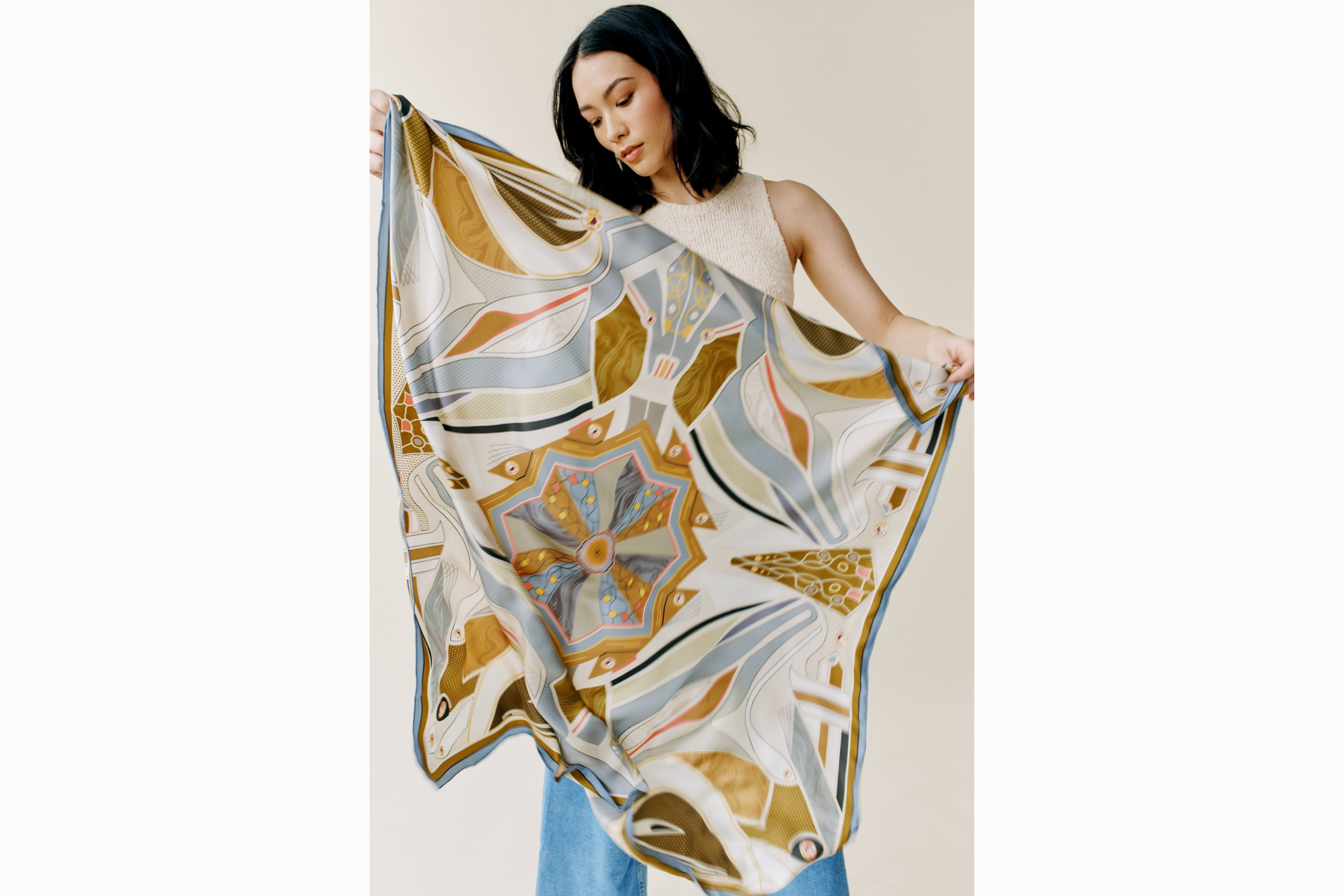 Model holds up 100% silk square scarf featuring rectilinear geometry, intricate line art, and aerodynamic curves in shades of cream and soft gray with pops of pink and metallic tones