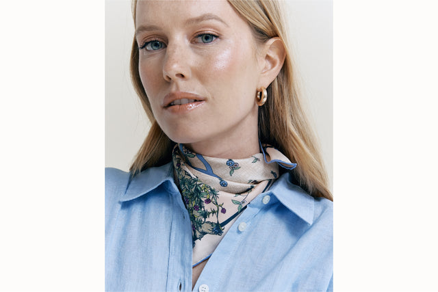 Image of a female model wearing the scarf wrapped around her and tied to be styled as a bandana along with a light blue button-down. 