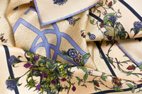 Close-up image of 100% silk square scarf featuring an English garden-inspired floral illustration motif with a periwinkle blue version of the brand logo dominantly featured. Illustrates the lightly ridged texture of the silk twill along with the rich color tones and luminous nature of the silk scarf. 