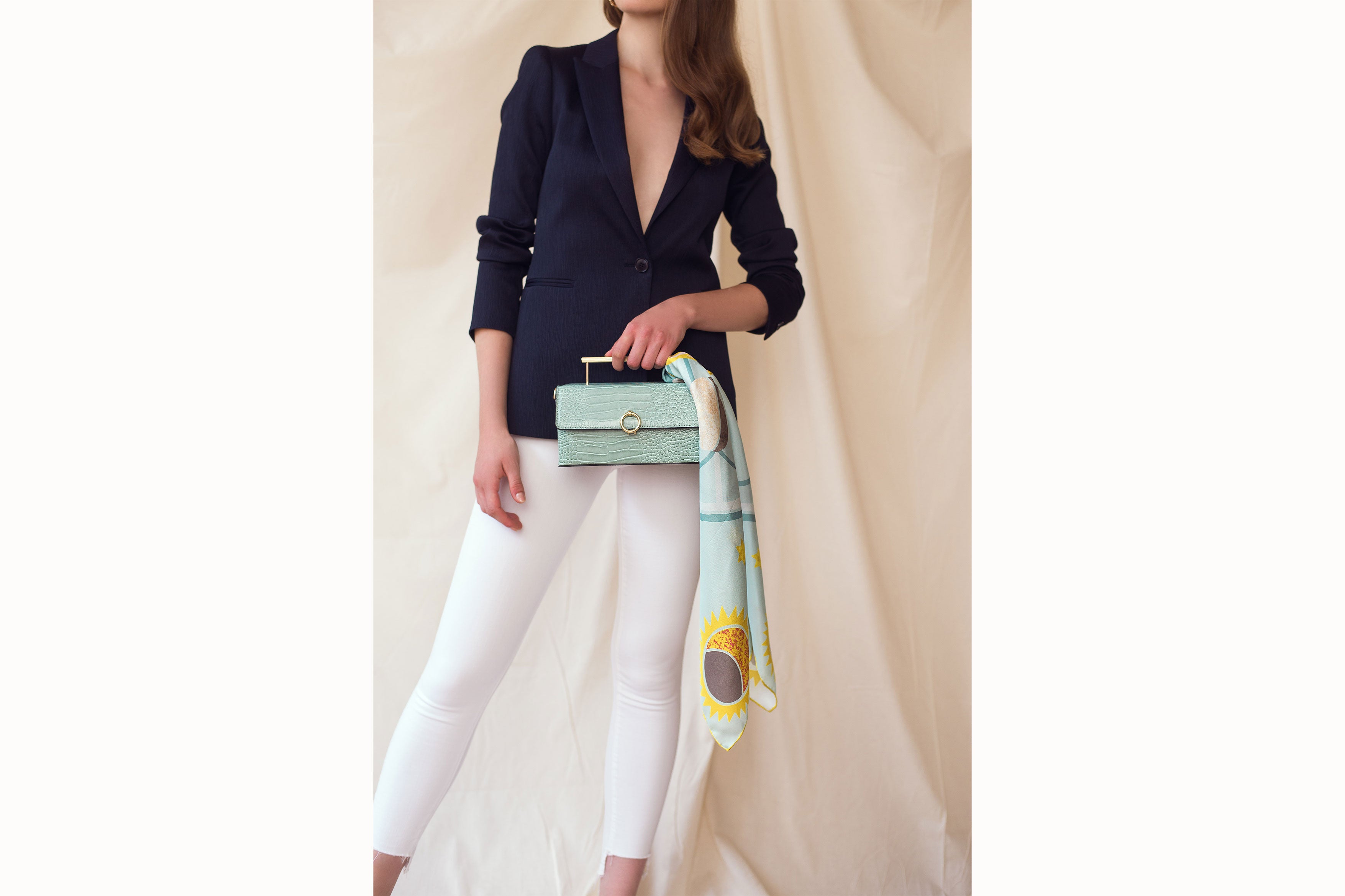 Image of female model wearing the scarf wrapped around the handle of a small turquoise leather handbag. Model is wearing white jeans and a navy blazer.