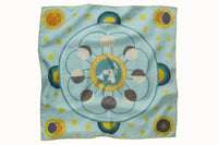 Flatlay image of 100% silk square scarf featuring a motif of stars, the sun and the moon phases positioned around planet Earth. Details in shades of rich turquoise, marine blue, white, chartreuse and maize on a light turquoise background. 