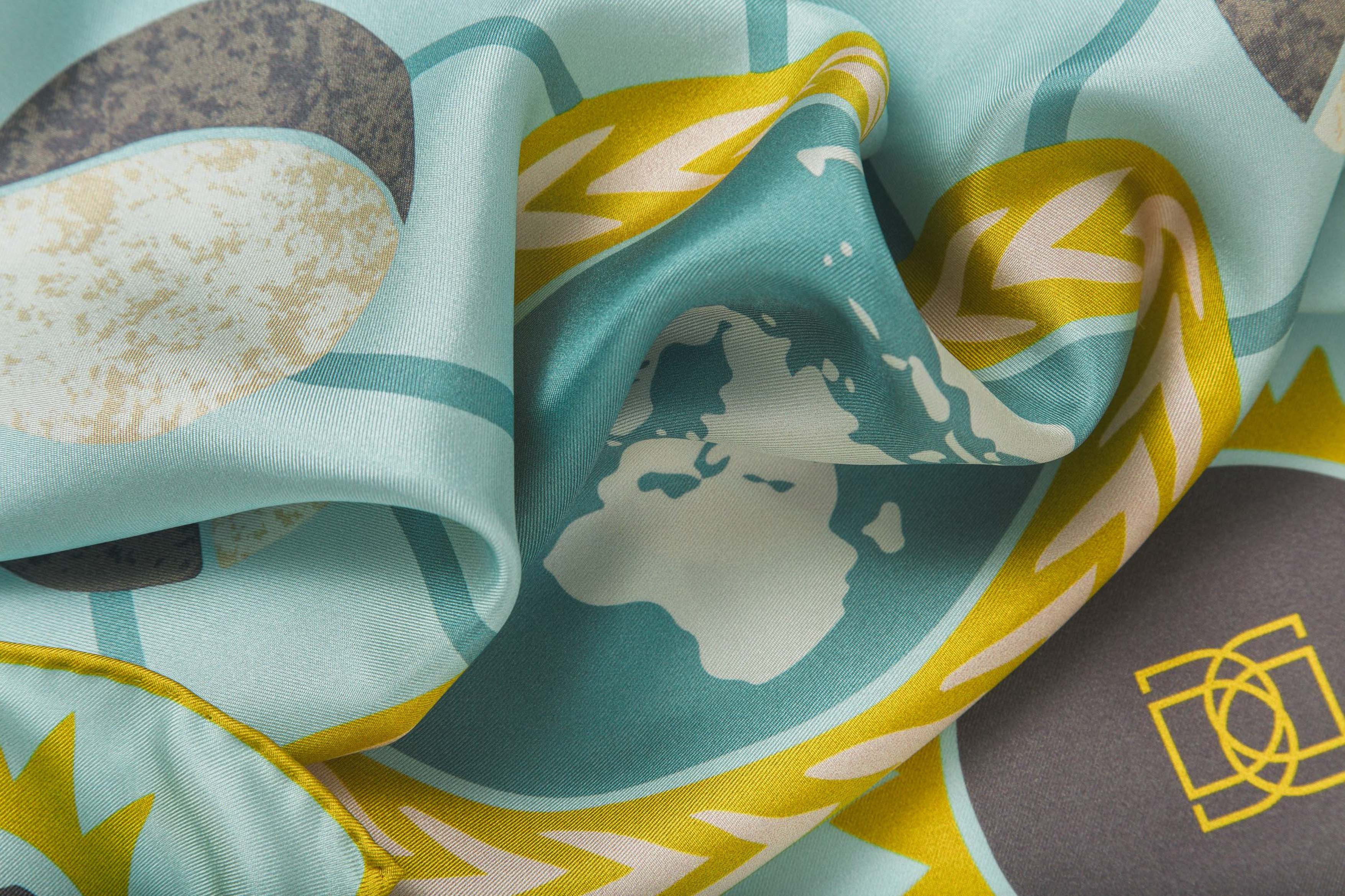 Close-up image of 100% silk square scarf featuring a motif of featuring a motif of stars, the sun and the moon phases positioned around planet Earth. Details in shades of rich turquoise, marine blue, white, chartreuse and maize on a light turquoise background. Illustrates the lightly ridged texture of the silk twill along with the rich color tones and luminous nature of the silk scarf.