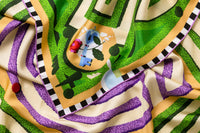 Close-up image of 100% silk square scarf featuring a motif of a garden labyrinth with green hedges and purple lavender elements on a warm cream background. Also features a blue key and floral detail in each of the four corners of the scarf as well as a black and white checker border around the edges. Illustrates the lightly ridged texture of the silk twill along with the rich color tones and luminous nature of the silk scarf.