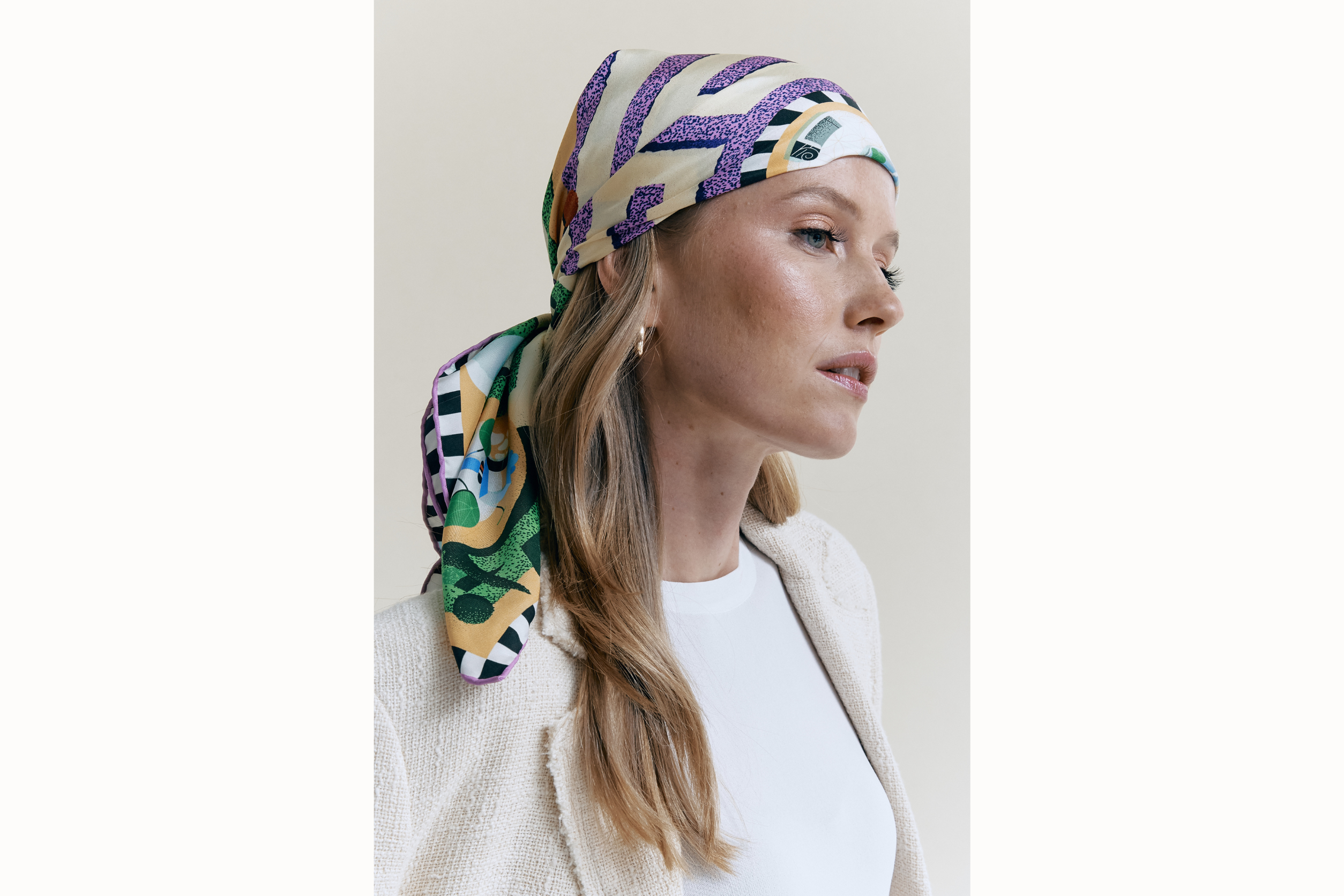 Image of a female model wearing a scarf wrapped around her head, tied in the back, styled as a headscarf. Model is wearing a white tank top and a cream blazer. 
