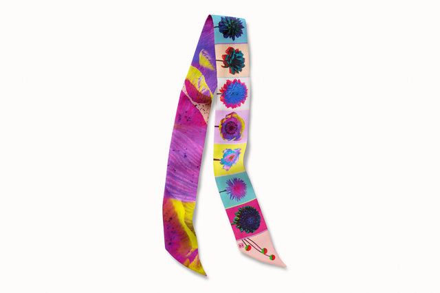 Flatlay image of 100% silk ribbon style scarf, 2” wide by 32” long, folded in half. Design features a series of brightly colored flowers in shades of pink, blue, green and yellow and an abstract close-up of a purple and yellow flower petal on the other.