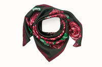 Rolled image of 100% silk square scarf featuring a motif of large-scale roses and leaves in shades of rich red and green on a background of deep forest green, with a dark burgundy border around the edges.