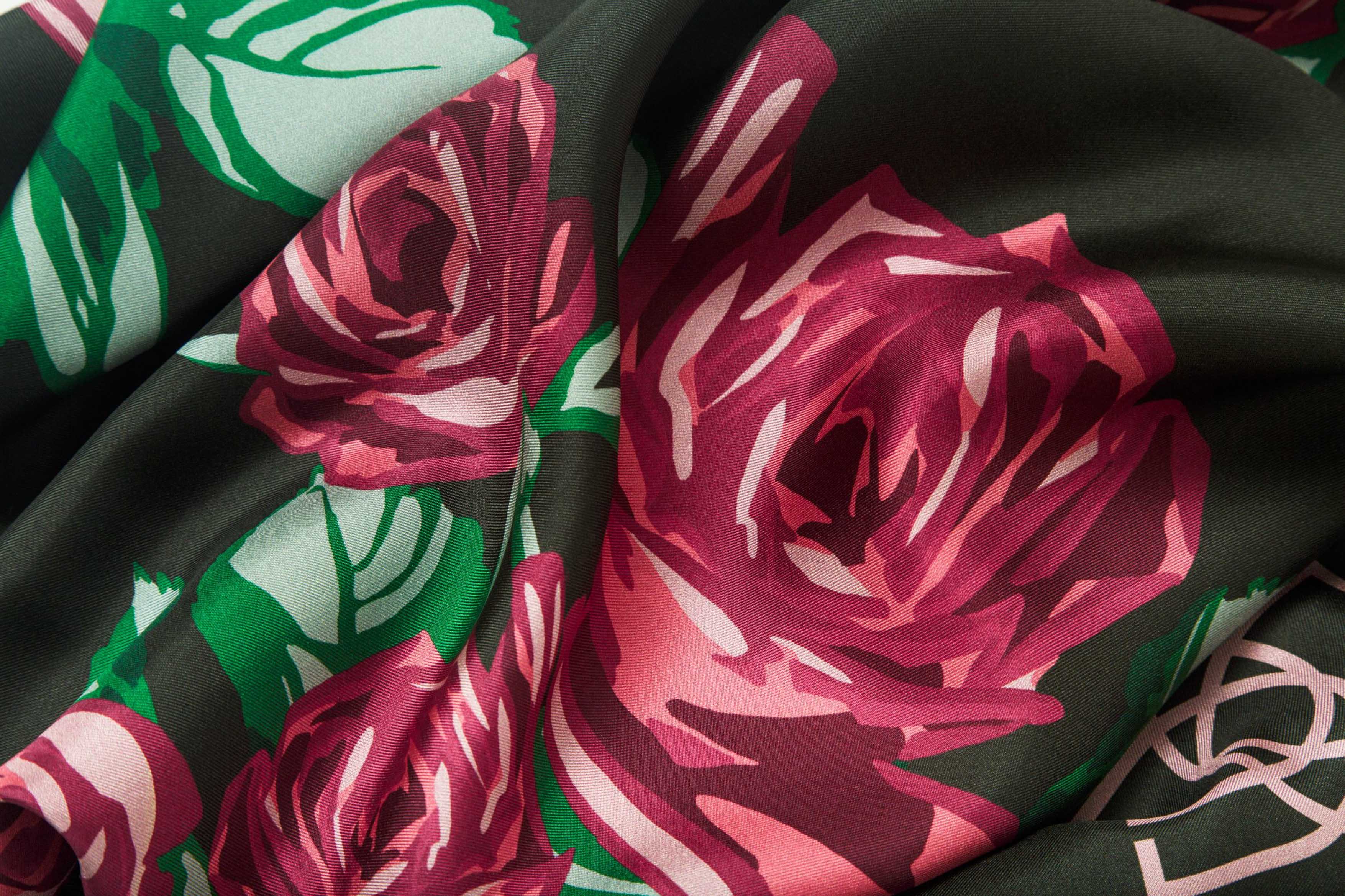 Close-up image of 100% silk square scarf featuring a motif of large-scale roses and leaves in shades of rich red and green on a background of deep forest green, with a dark burgundy border around the edges. Illustrates the lightly ridged texture of the silk twill along with the rich color tones and luminous nature of the silk scarf.