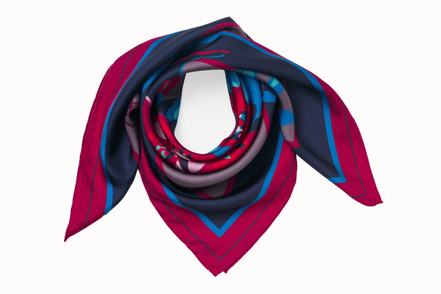 Rolled image of 100% silk square scarf featuring a motif of large-scale florals and leaves in shades of rich red, magenta, berry and marine blues on a background of smoky lilac, with a dark blue and magenta border around the edges.