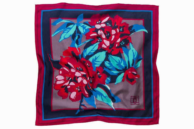 Flatlay image of 100% silk square scarf featuring a motif of large-scale florals and leaves in shades of rich red, magenta, berry and marine blues on a background of smoky lilac, with a dark blue and magenta border around the edges.