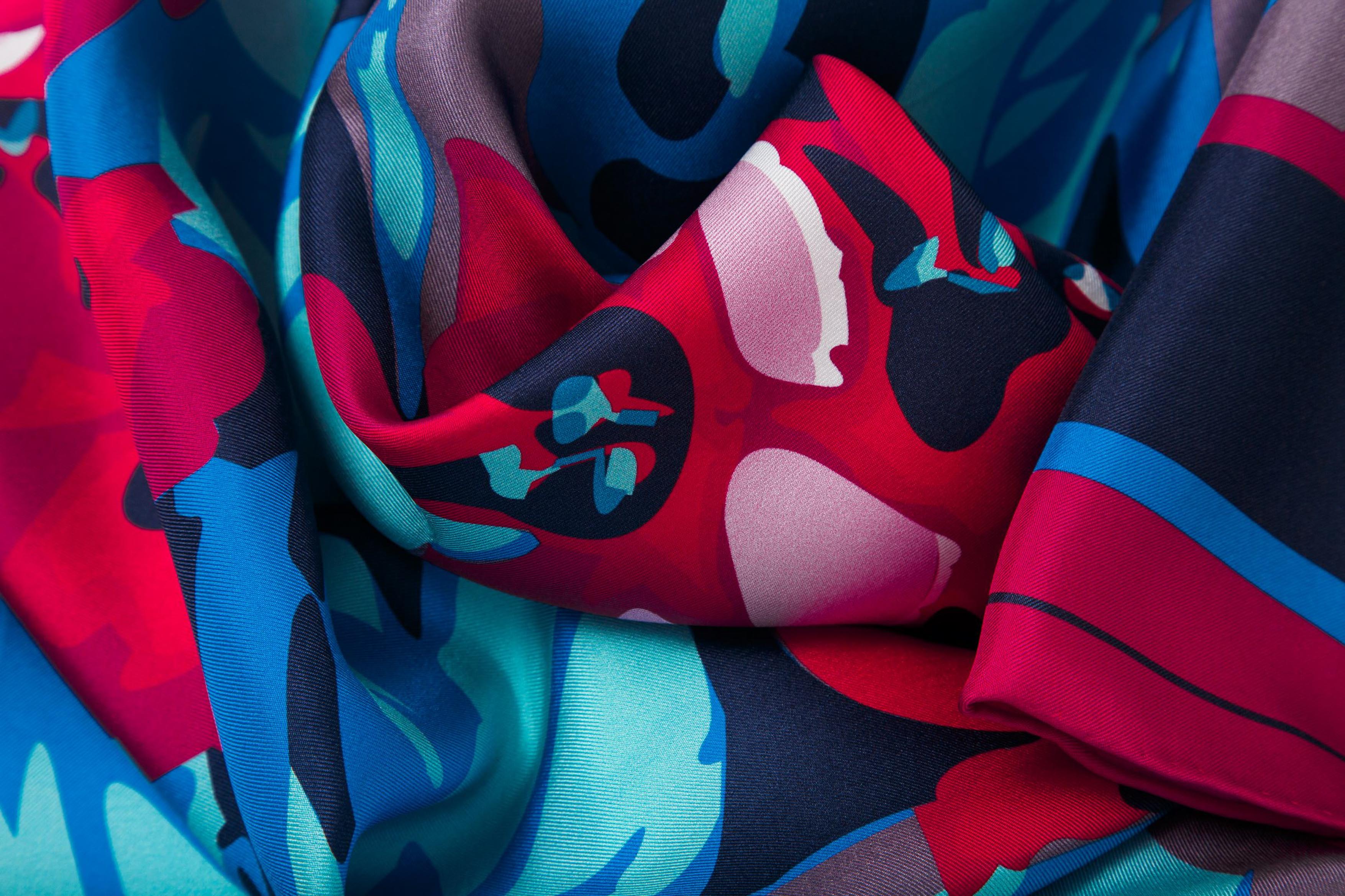 Close-up image of 100% silk square scarf featuring a motif of large-scale florals and leaves in shades of rich red, magenta, berry and marine blues on a background of smoky lilac, with a dark blue and magenta border around the edges. Illustrates the lightly ridged texture of the silk twill along with the rich color tones and luminous nature of the silk scarf.