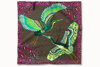 Flatlay image of 100% silk square scarf featuring a motif of two large-scale cranes with brightly colored wings in shades of blue and green with geometric designs. The scarf’s center background is a tonal army green and the border features irregular grey polka dots on a magenta background.