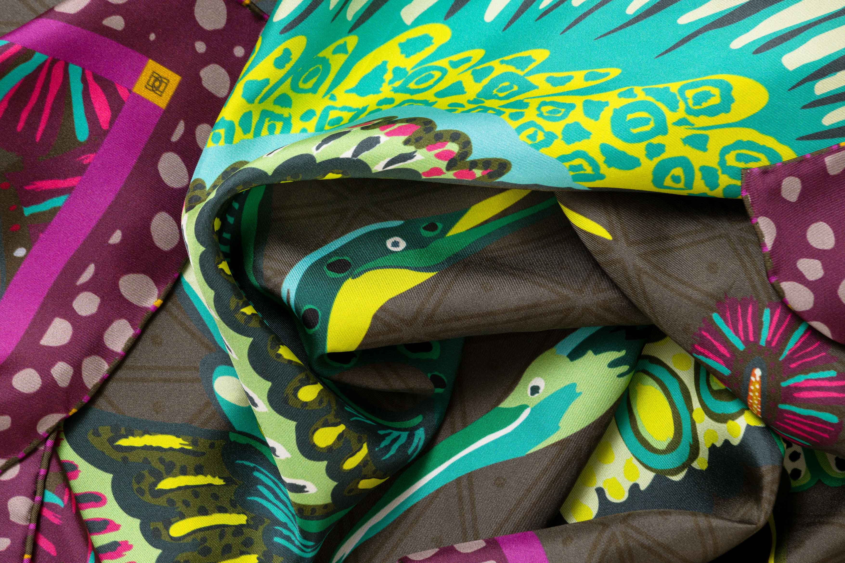 Close-up image of 100% silk square scarf featuring a motif of two large-scale cranes with brightly colored wings in shades of blue and green with geometric designs. The scarf’s center background is a tonal army green and the border features irregular grey polka dots on a magenta background. Illustrates the lightly ridged texture of the silk twill along with the rich color tones and luminous nature of the silk scarf.
