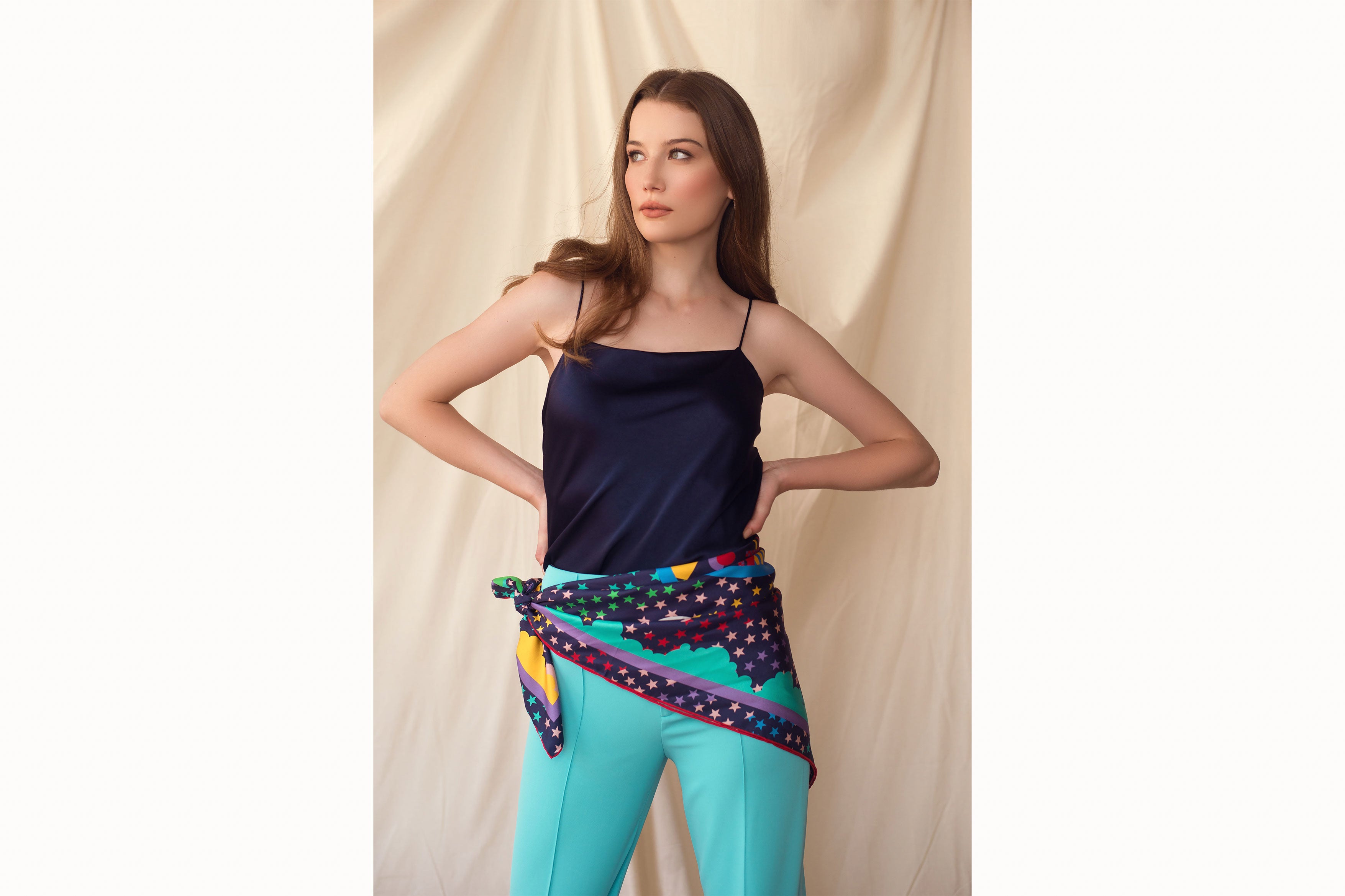 Image of female model standing with hands on hips while wearing the scarf tied around her waist. Model is also wearing a navy tank top and turquoise trousers.