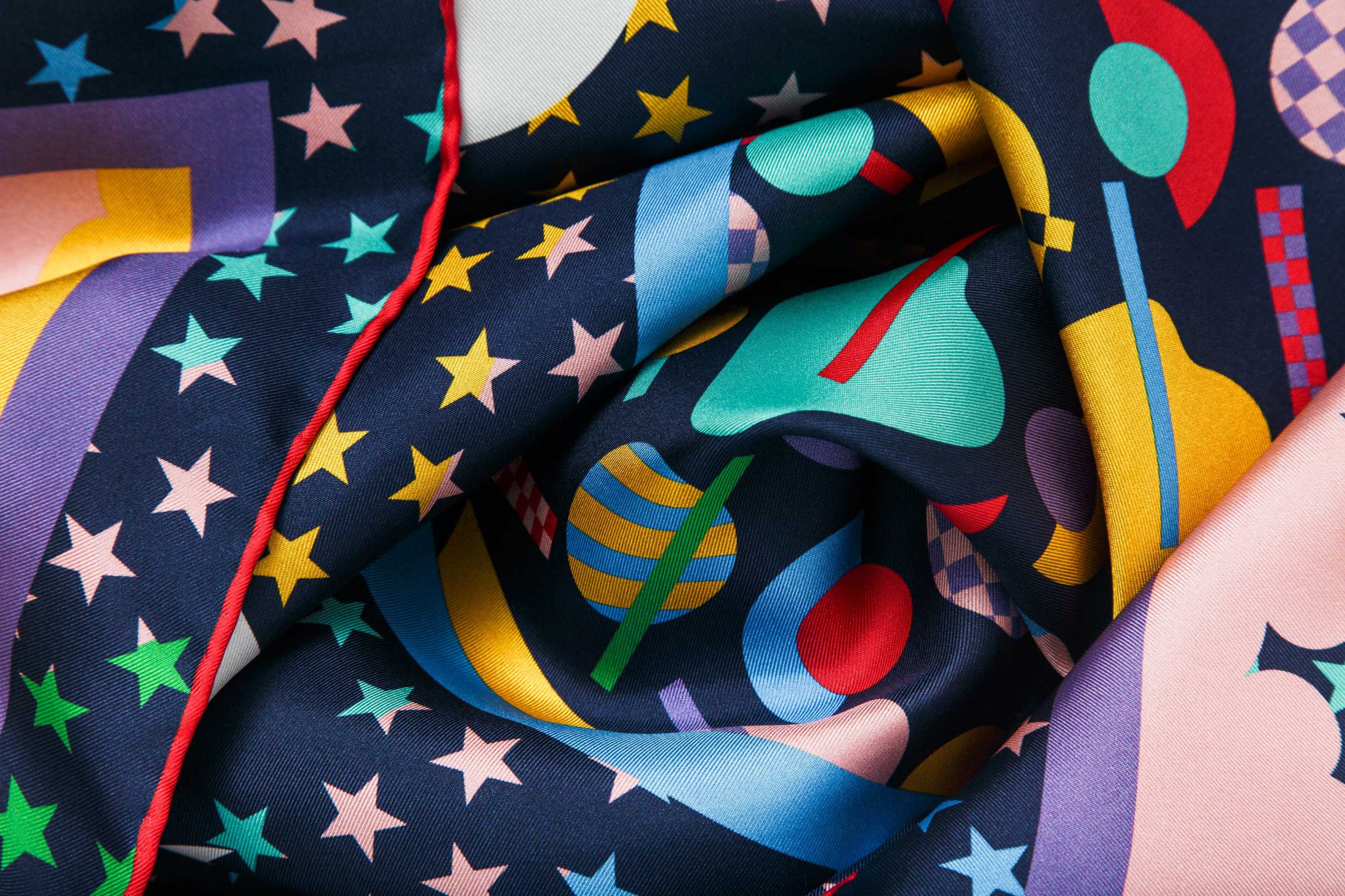 Close-up image of 100% silk square scarf featuring a motif of brightly colored stars, clouds and white moon phases throughout. The center features colorful geometric shapes as a compliment to the celestial theme. Illustrates the lightly ridged texture of the silk twill along with the rich color tones and luminous nature of the silk scarf.