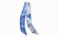 Flatlay image of 100% silk ribbon style scarf, 2” wide by 32” long. Featuring a illustrations of Greek goddess busts and urns in a bright Aegean blue on a white background. The opposite side of the scarf features a white Greek key design motif on a bright blue background.