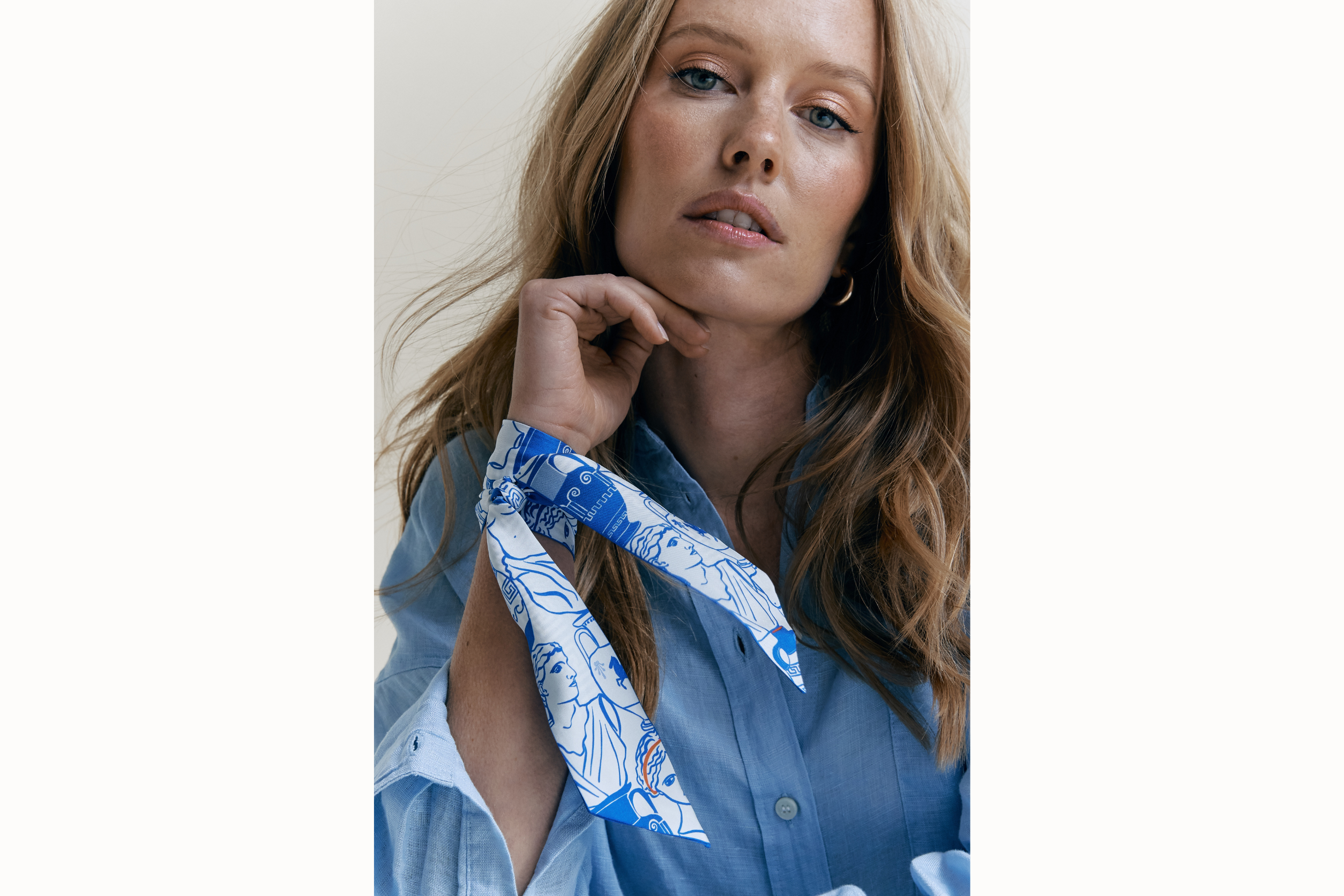  Image of a female model wearing the scarf wrapped around her neck with tail ends of the scarf draping onto her arm. Model is wearing a light blue button down and her hair is brushed to the right side of her body.