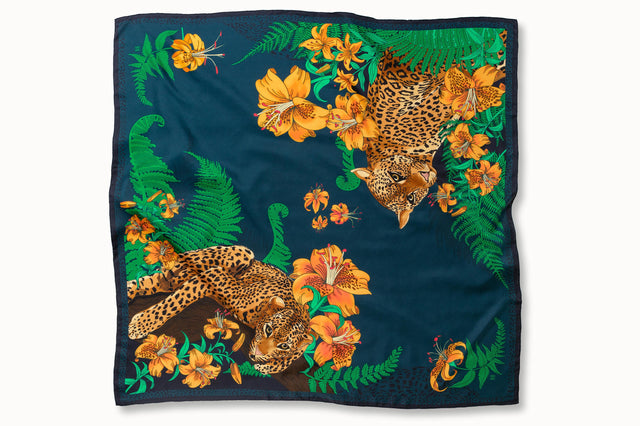 Flatlay image of 100% silk square scarf featuring a motif of two leopards lounging in a jungle scene surrounded by lush greenery and tiger lily florals on a marine blue background.