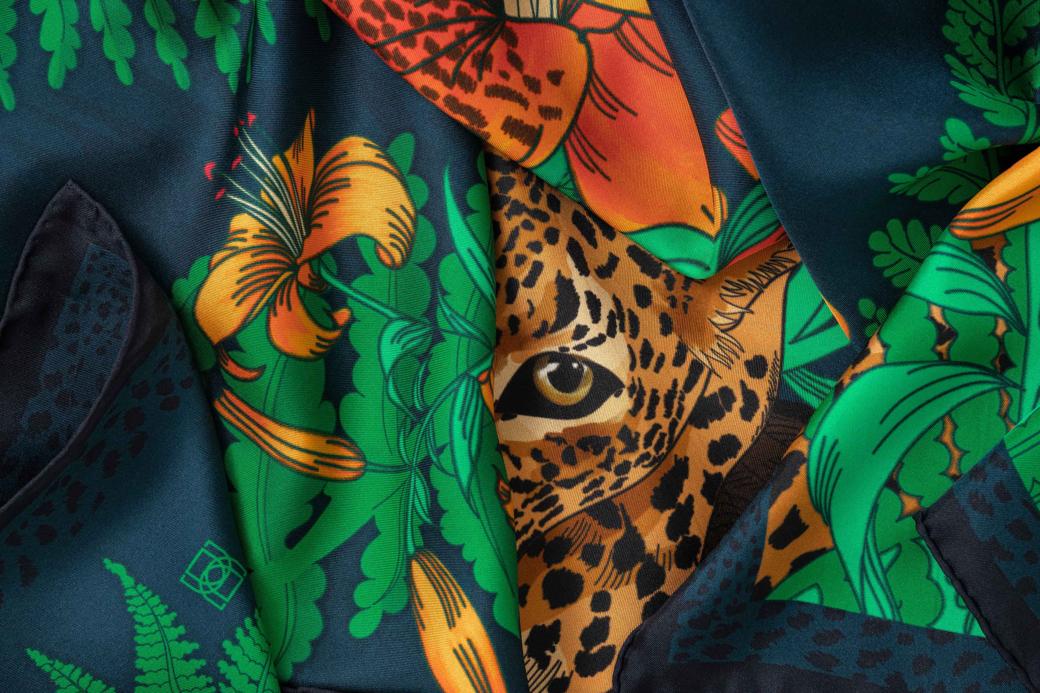 Close-up image of 100% silk square scarf featuring a motif of two leopards lounging in a jungle scene surrounded by lush greenery and tiger lily florals on a marine blue background. Illustrates the lightly ridged texture of the silk twill along with the rich color tones and luminous nature of the silk scarf.