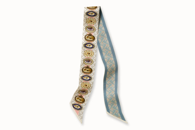 Flatlay image of 100% silk ribbon style scarf, 2” wide by 32” long, folded in half. Design features a series of hand-painted lover's eye jewels on one side and hand drawn wallpaper design with a floral motif in a soft blue tone on the other side.