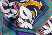 Close-up image of 100% silk square scarf featuring a motif of black and white intertwined hands. The larger scale hands motif in the center is on a pumpkin colored background and is surrounded by smaller scale hands on a pink coral colored background. The scarf has a green and purple border. Illustrates the lightly ridged texture of the silk twill along with the rich color tones and luminous nature of the silk scarf.