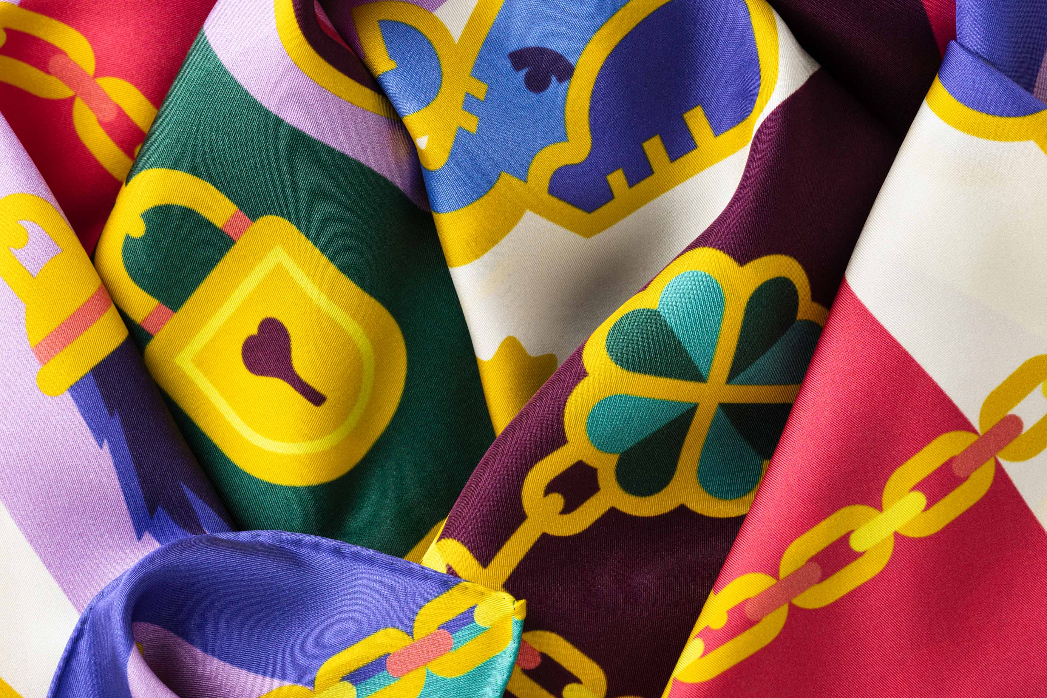 Close-up image of 100% silk square scarf featuring a motif of jewel toned good luck charms such as ladybugs, horseshoes, four leaf clovers, wishbones on a large gold chain with the evil eye symbol at the center. Illustrates the lightly ridged texture of the silk twill along with the rich color tones and luminous nature of the silk scarf.]